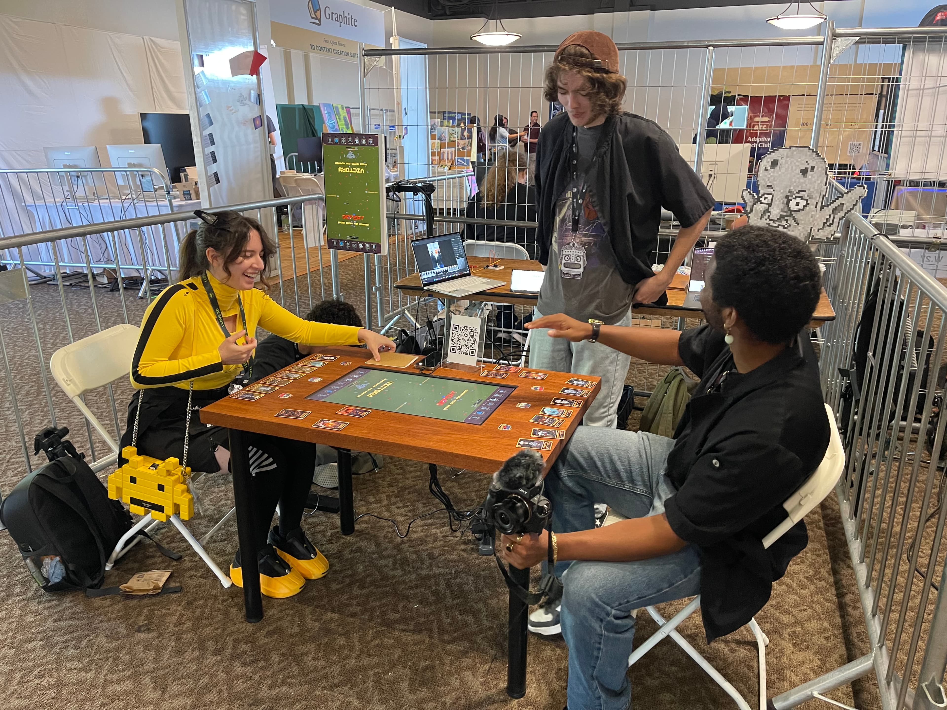A group playing UVULITES around the finished table at an event booth — cards dealt around the embedded screen while onlookers watch and film