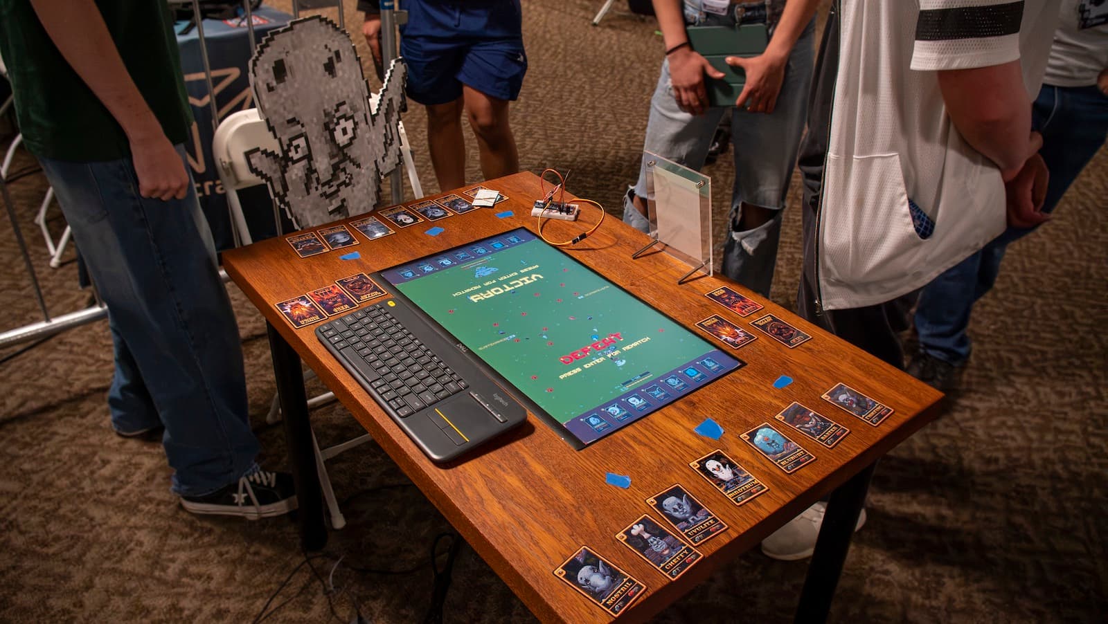 Side view of the finished UVULITES table mid-match at an event — embedded screen in the wooden top with NFC cards placed along both reader edges